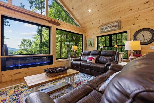 A cozy living room with a leather sofa, a fireplace, and large windows offering a view of the outdoors.