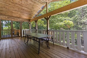 An outdoor wooden deck with a table and chairs, surrounded by a white railing, overlooking a forested area.