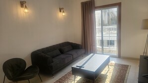 A living room with a dark sofa, a small table, a chair, and a view of a balcony through a sliding door. - 106 NIC - Stay Kallisto Old Town Apartment (Nicosia)