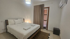 A neatly made bed with white linens and a folded towel, a bedside lamp, a window with blinds, and a wall-mounted air conditioner. - 106 NIC - Stay Kallisto Old Town Apartment (Nicosia)