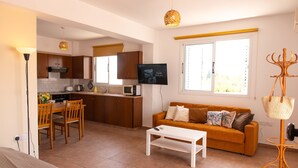 Ein modernes Wohnzimmer mit einer Couch, einem Couchtisch und einem Fernseher. Es gibt eine Küchenzeile mit Holzschränken und einem Esstisch mit Stühlen.