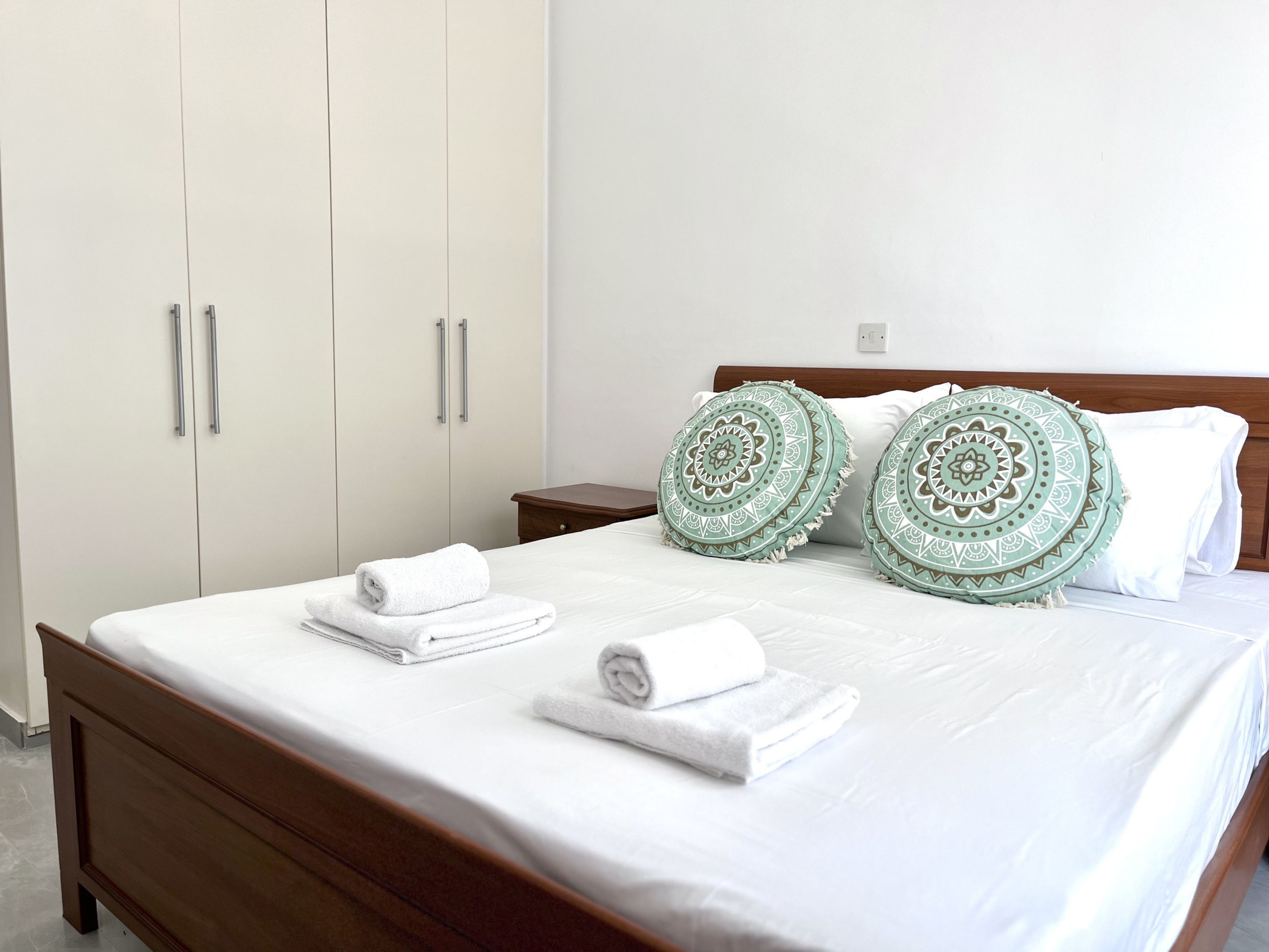 A neatly made bed with white bedding and two decorative pillows, a wooden nightstand, and a wardrobe with silver handles.