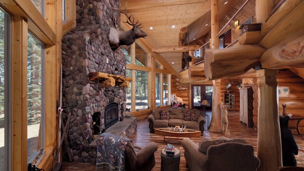 A log cabin interior with a stone fireplace, wooden furniture, and elk antlers mounted on the wall. - Little Deschutes Wildlife Lodge - Nature's Cabin (La Pine)