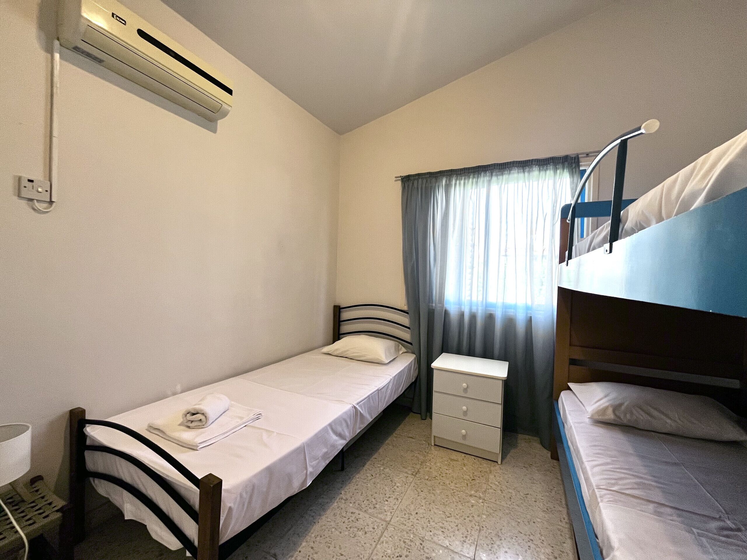 A small, single-bed room with a bunk bed, a bedside table, an air conditioning unit, and a window with curtains.