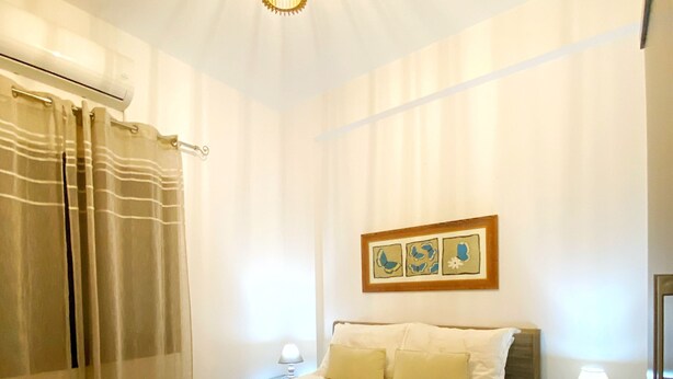 Una cama bien hecha con cubrecama acolchado, dos lámparas de noche, un cuadro en la pared y una ventana con cortinas.