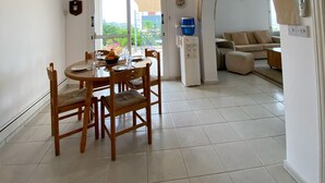 A spacious dining area with a round table and chairs, a water dispenser, and a view through a glass door to a balcony. - 102 PAP - Stay Helios House (Mesa Chorio)