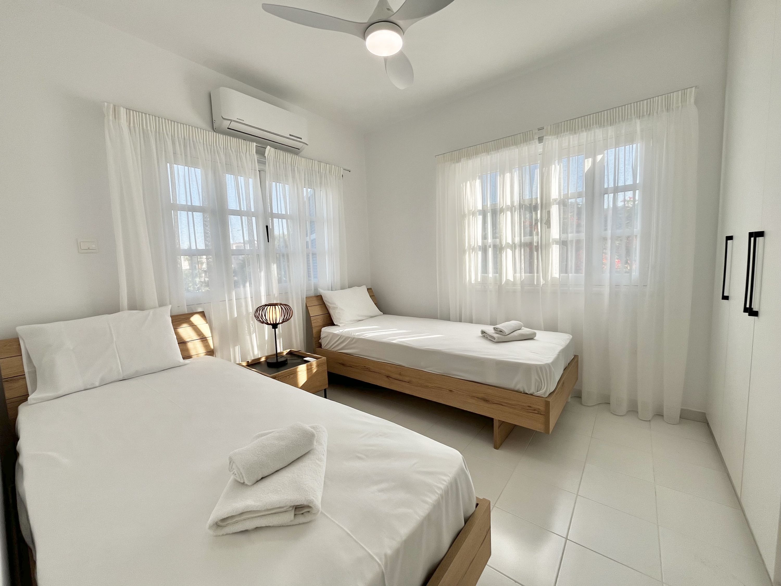 A hotel room with two beds, a ceiling fan, an air conditioning unit, and large windows with sheer curtains.
