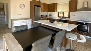 A modern kitchen with a central island, wooden cabinets, and a dining area with a black table and chairs. - 121 PEY - Stay Poseidon Villa (Pegeia)