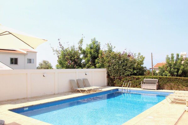 Piscina con sillones de descanso y una sombrilla, rodeada por un muro blanco y vegetación.