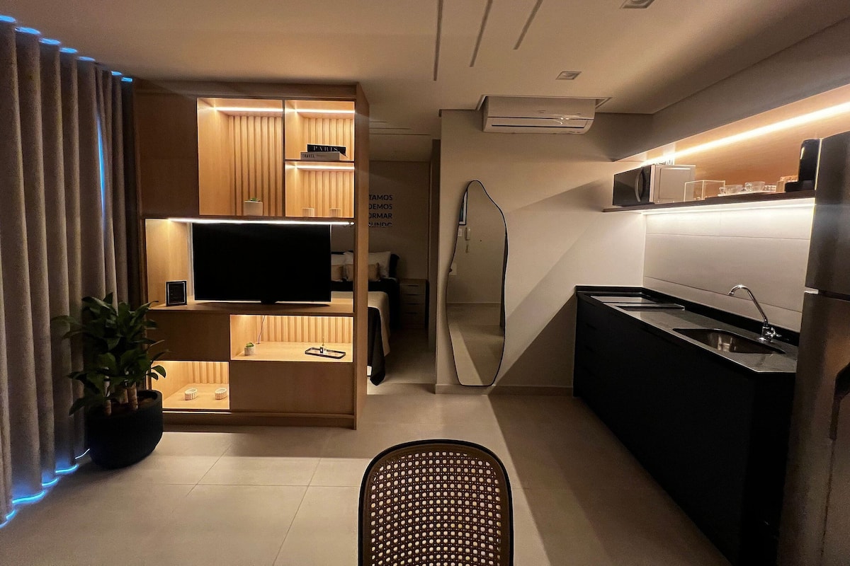 A modern kitchen with a built-in microwave, a refrigerator, and a sink. There is a TV on a wooden shelf, and a potted plant is visible.