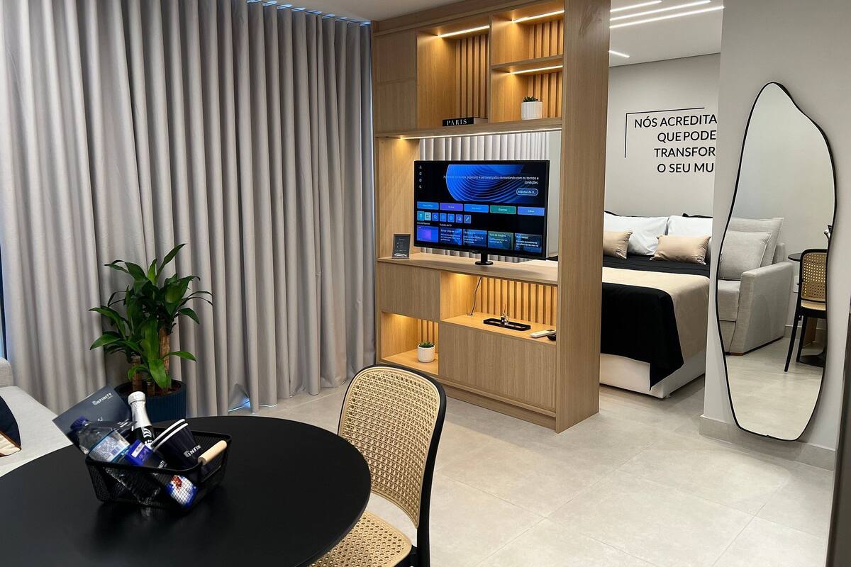 A modern hotel room with a flat-screen TV, a bed, a seating area with a table and chairs, and a large mirror.
