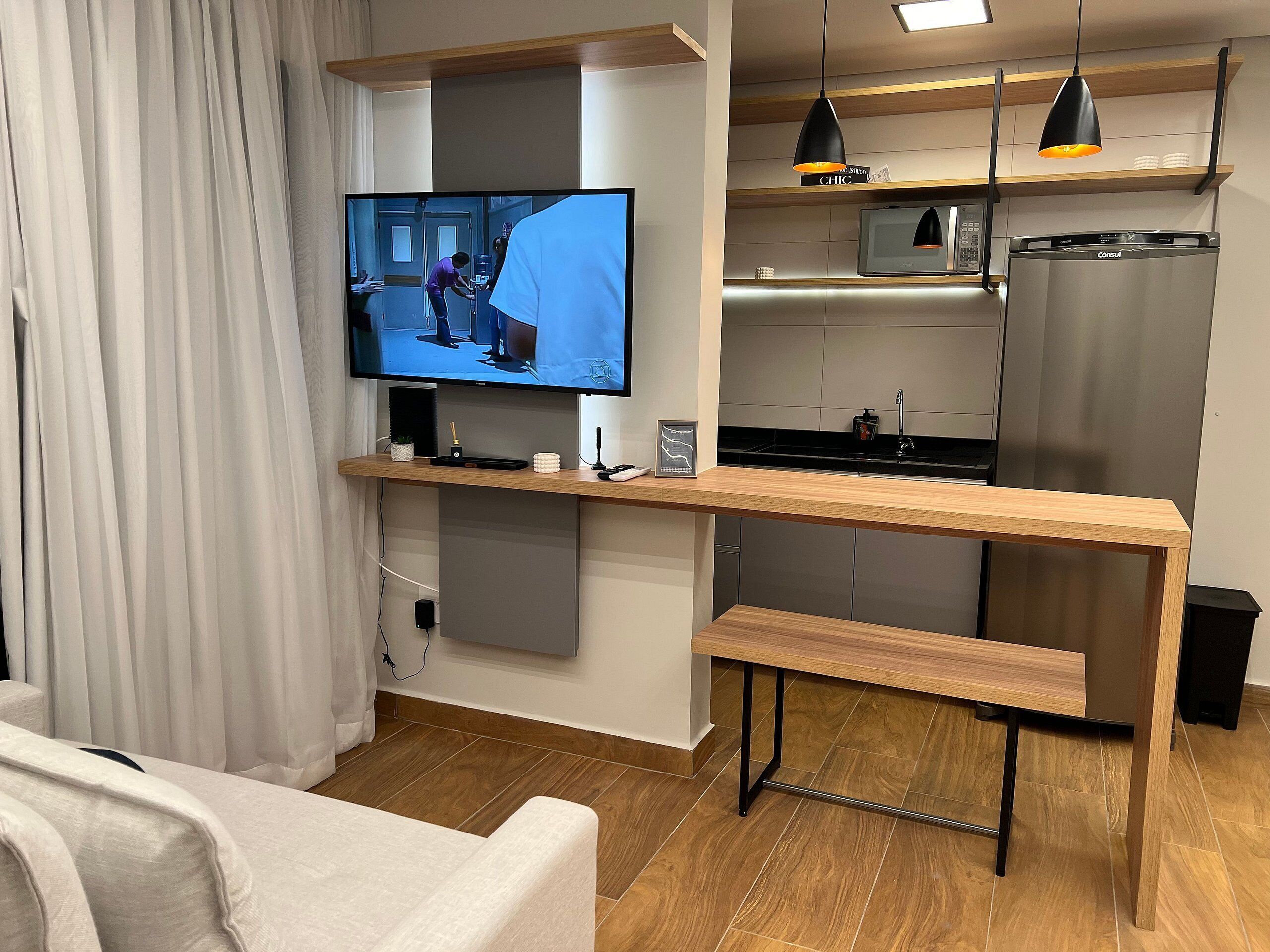 A modern kitchen with a built-in oven, microwave, and refrigerator. A wooden bar counter with a bench is in front of the kitchen, and a flat-screen TV is mounted above it.