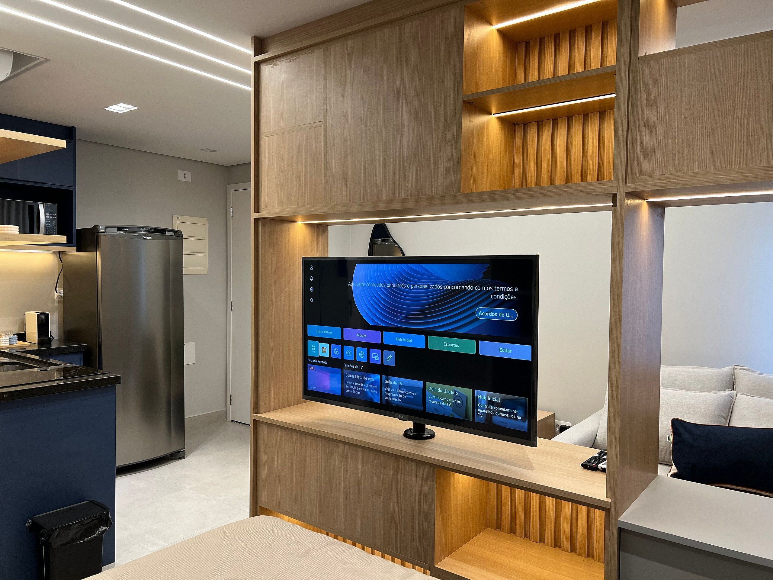 A modern kitchen with a built-in refrigerator, a flat-screen TV mounted on a wooden wall, and a seating area with a couch and pillows.