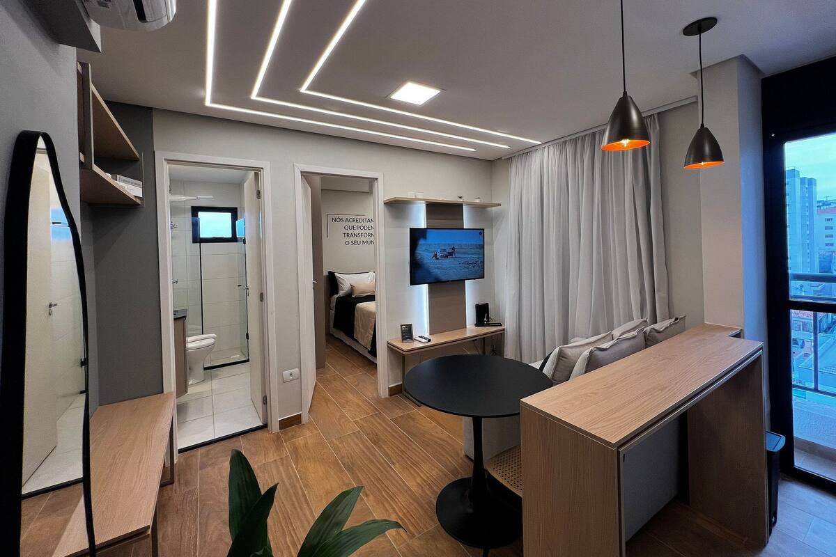 A modern hotel room with a wooden floor, a desk, a TV, and a large window with curtains.