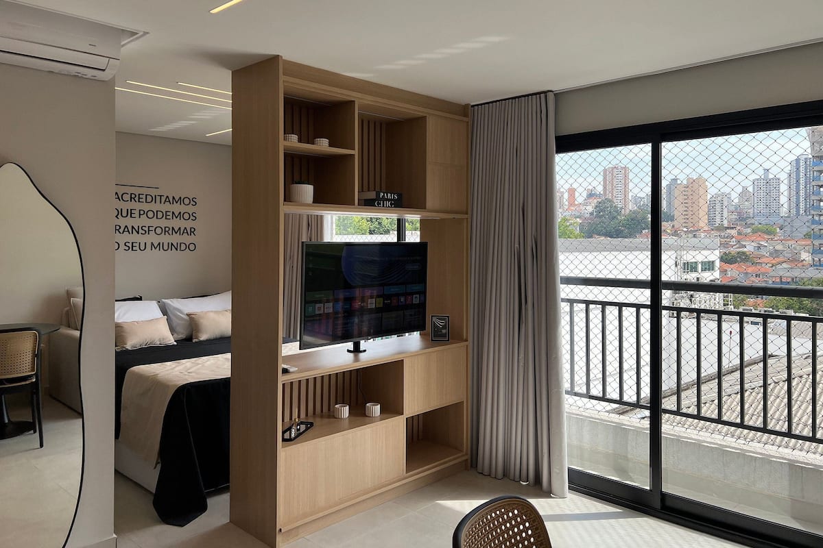 A modern hotel room with a large TV, a bed, a seating area, and a balcony with a city view.