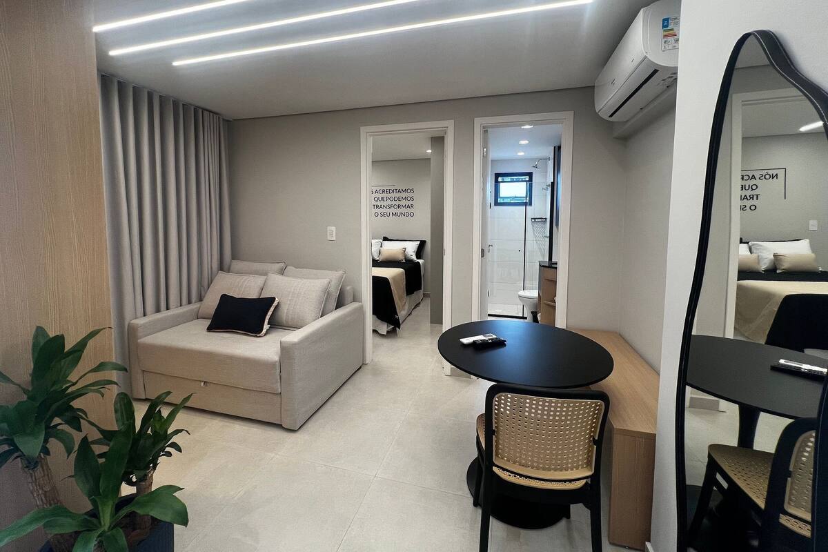 A modern, well-lit room with a sofa, a dining table, and a chair. There is a mirror on one wall and an air conditioning unit above it.