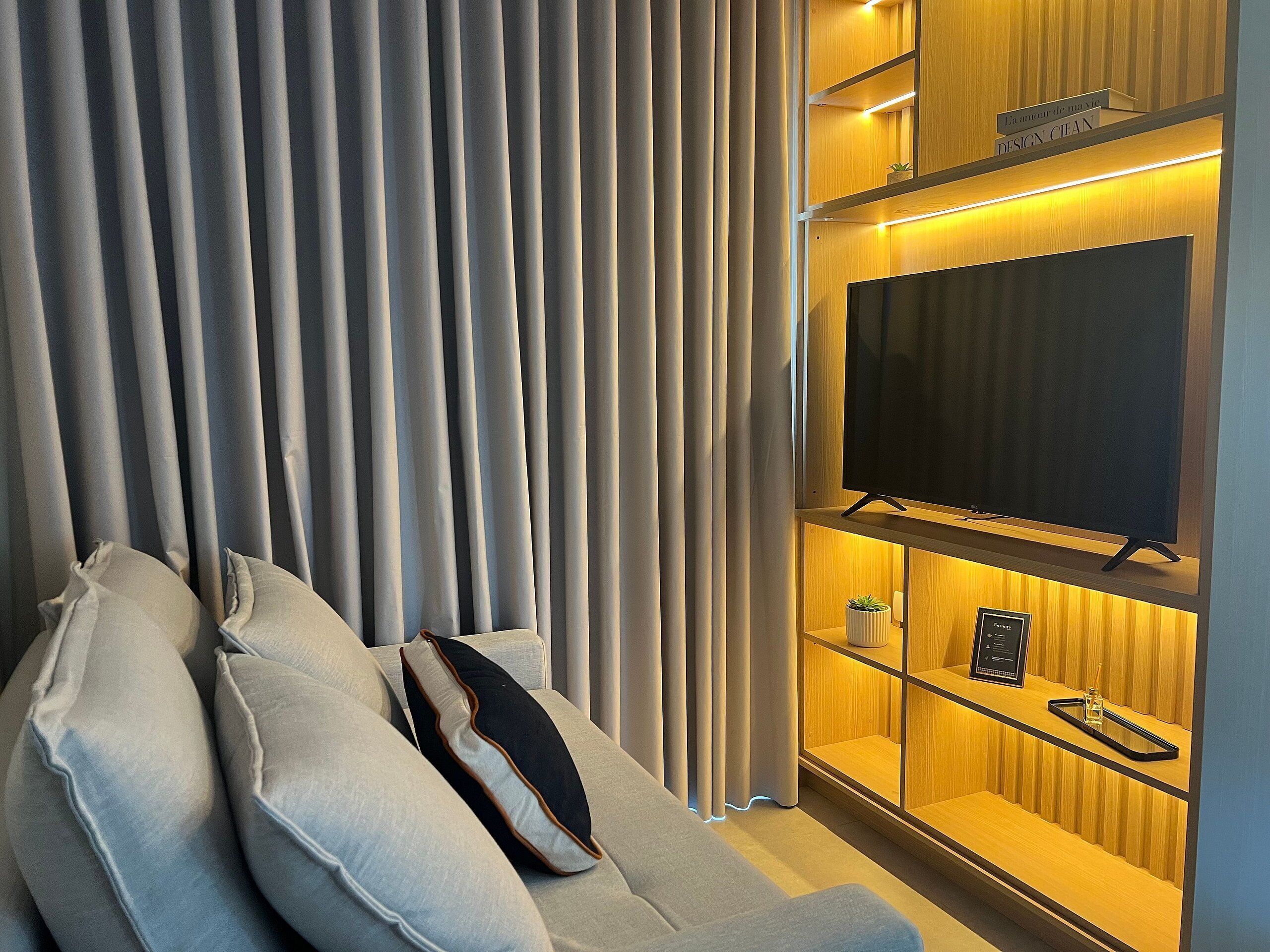 A modern living room with a sofa, a flat-screen TV mounted on a wooden shelf, and a set of vertical blinds.