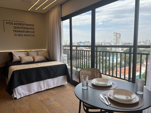 A hotel room with a large bed, a balcony with a city view, and a dining area with plates and cutlery. - Infinity Home Suítes Anhembi (Sao Paulo)