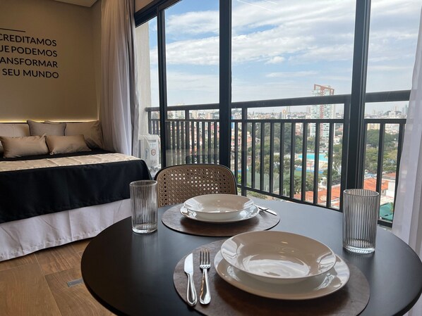 Ett hotellrum med en balkong, en säng med ett svartvitt sängöverkast, ett matbord dukat med tallrikar, bestick och glas, och utsikt över stadsbilden.