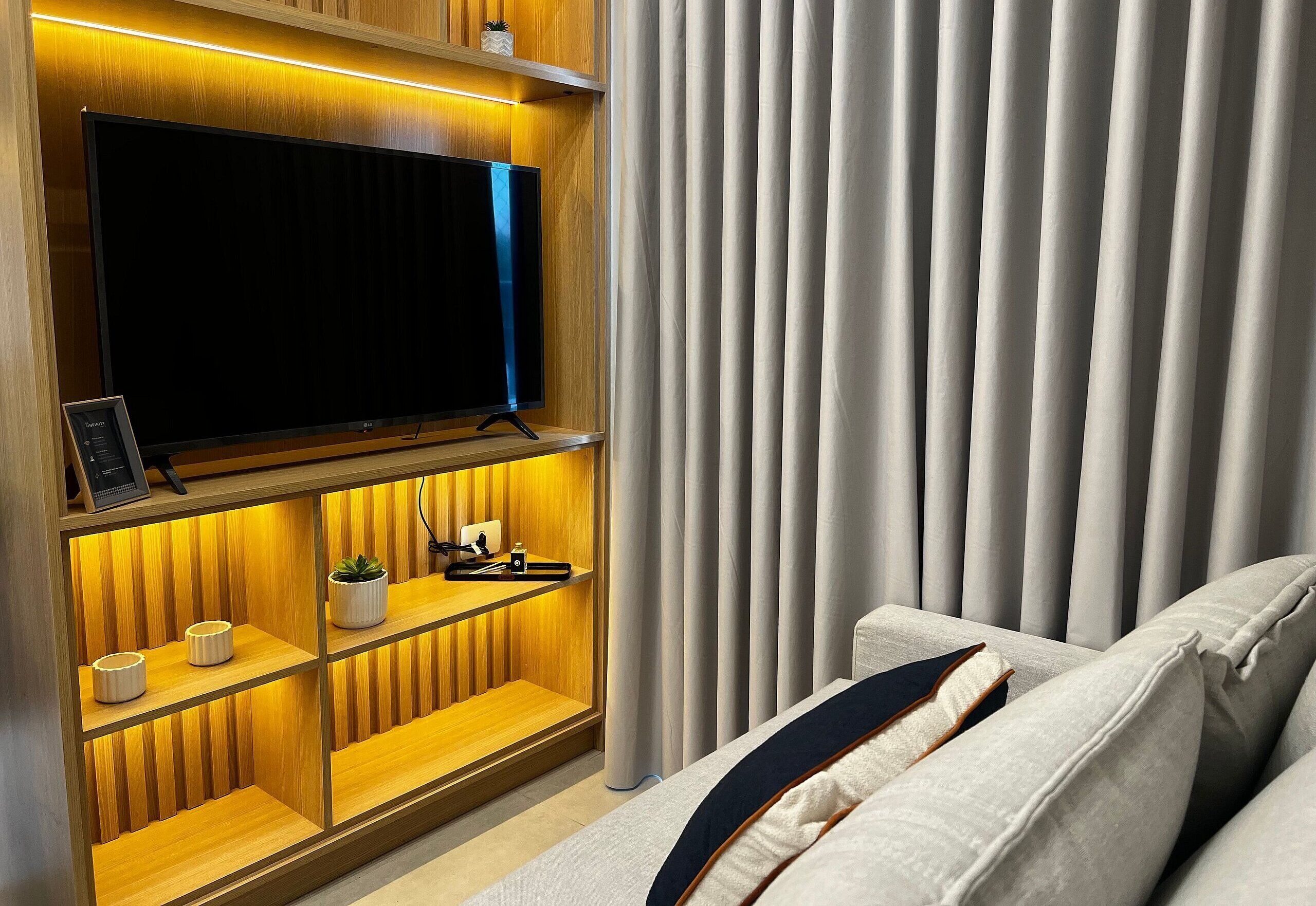 Ein modernes Wohnzimmer mit einem an einer Holzwand montierten Flachbildfernseher, einem Sofa mit dunklen Kissen und einem Vorhang auf der rechten Seite.