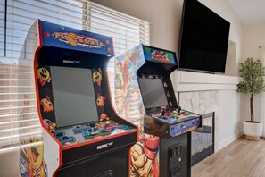 Duas máquinas de jogos com a marca 'Arcade Up', uma televisão e um vaso de planta num quarto com lareira.