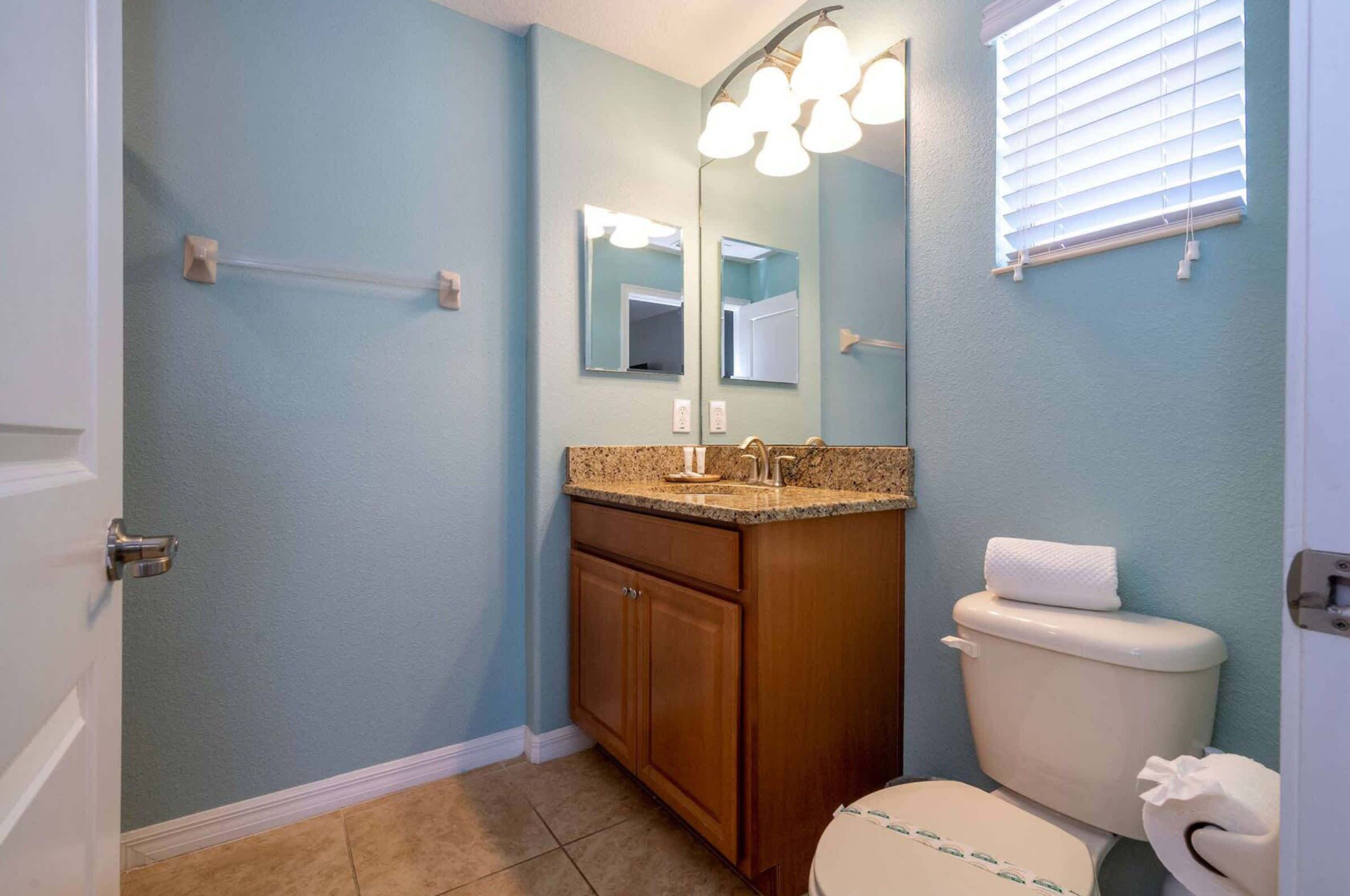 A bathroom with a blue wall, a wooden vanity, a white toilet, and a mirror with lights above it.