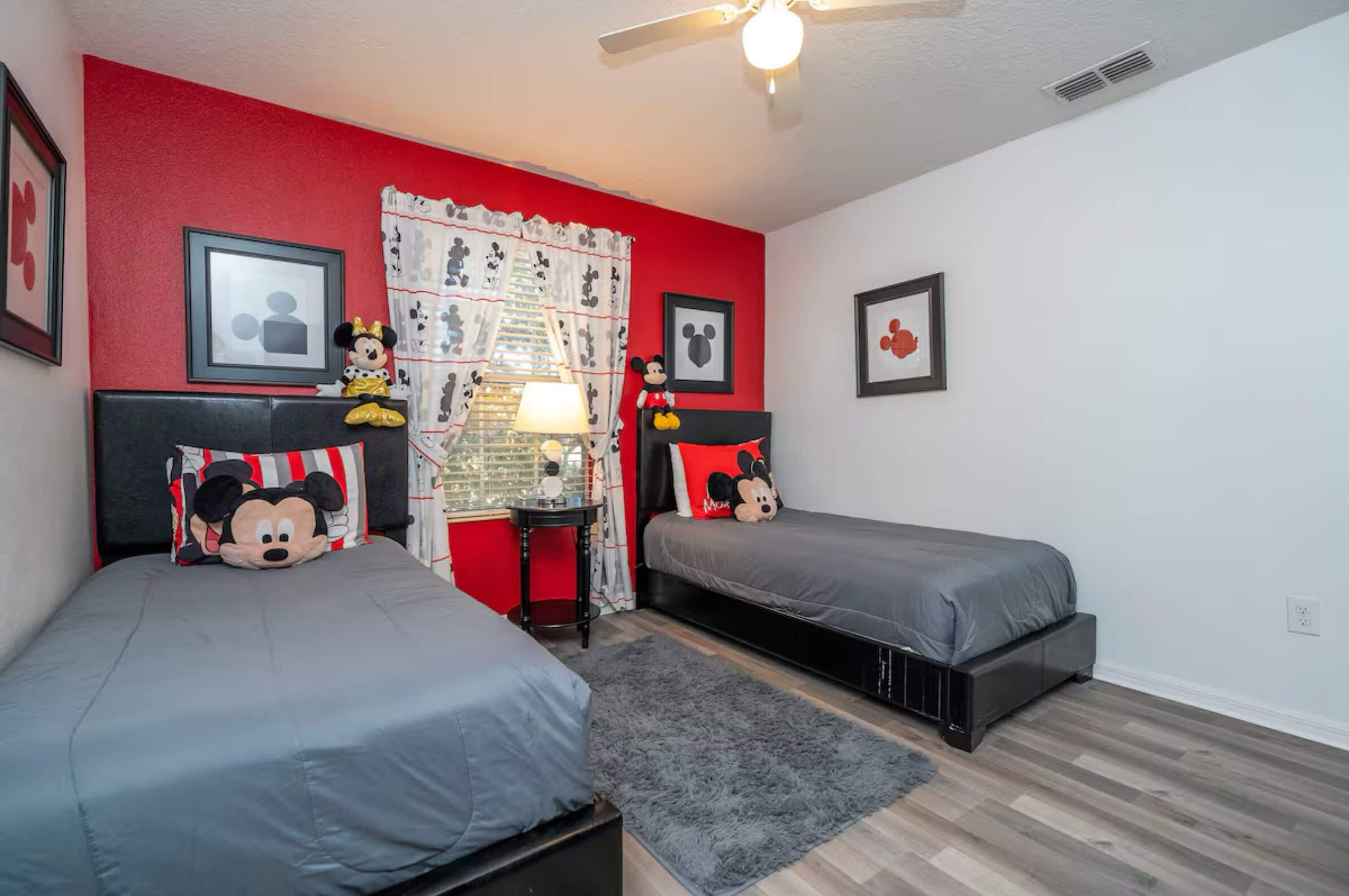 Quarto com duas camas, paredes vermelhas, almofadas do Mickey Mouse, uma mesinha com abajur e um ventilador de teto.