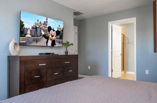 A bedroom with a large bed, a dresser with drawers, a flat-screen TV on the wall, and a door leading to another room.
