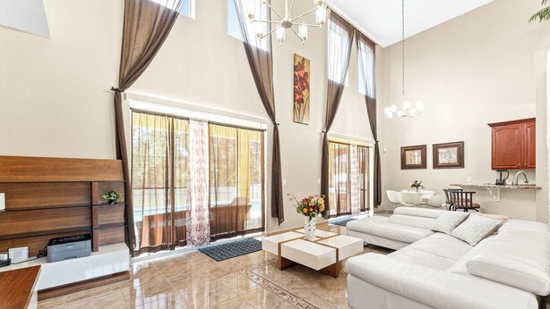A spacious living room with a large sofa, a coffee table, and a fireplace. There are large windows with curtains, a chandelier, and a kitchen area in the background. - Jasmine Breeze Home for 12 (Kissimmee)