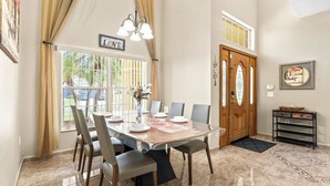 A dining area with a table set for six, a chandelier, and a view of the outdoors through large windows. - Jasmine Breeze Home for 12 (Kissimmee)