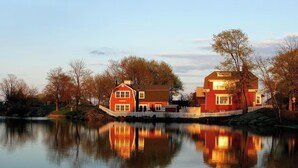 Een groot, rood, twee verdiepingen tellend huis met een witte hekken, gelegen aan een rustig waterlichaam dat het huis en de bomen weerspiegelt.