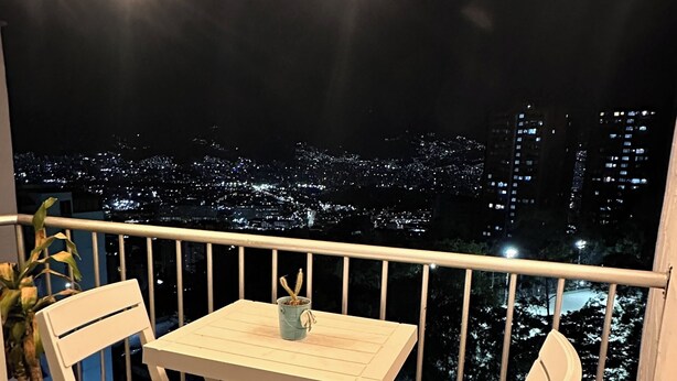 Un balcon avec une table et des chaises, des guirlandes lumineuses et une vue sur la ville la nuit.