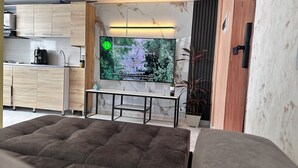 Un salon moderne doté d’un grand téléviseur à écran plat fixé au mur, d’un meuble en bois, d’une petite table avec un verre à vin et d’un coin salon confortable avec des coussins.