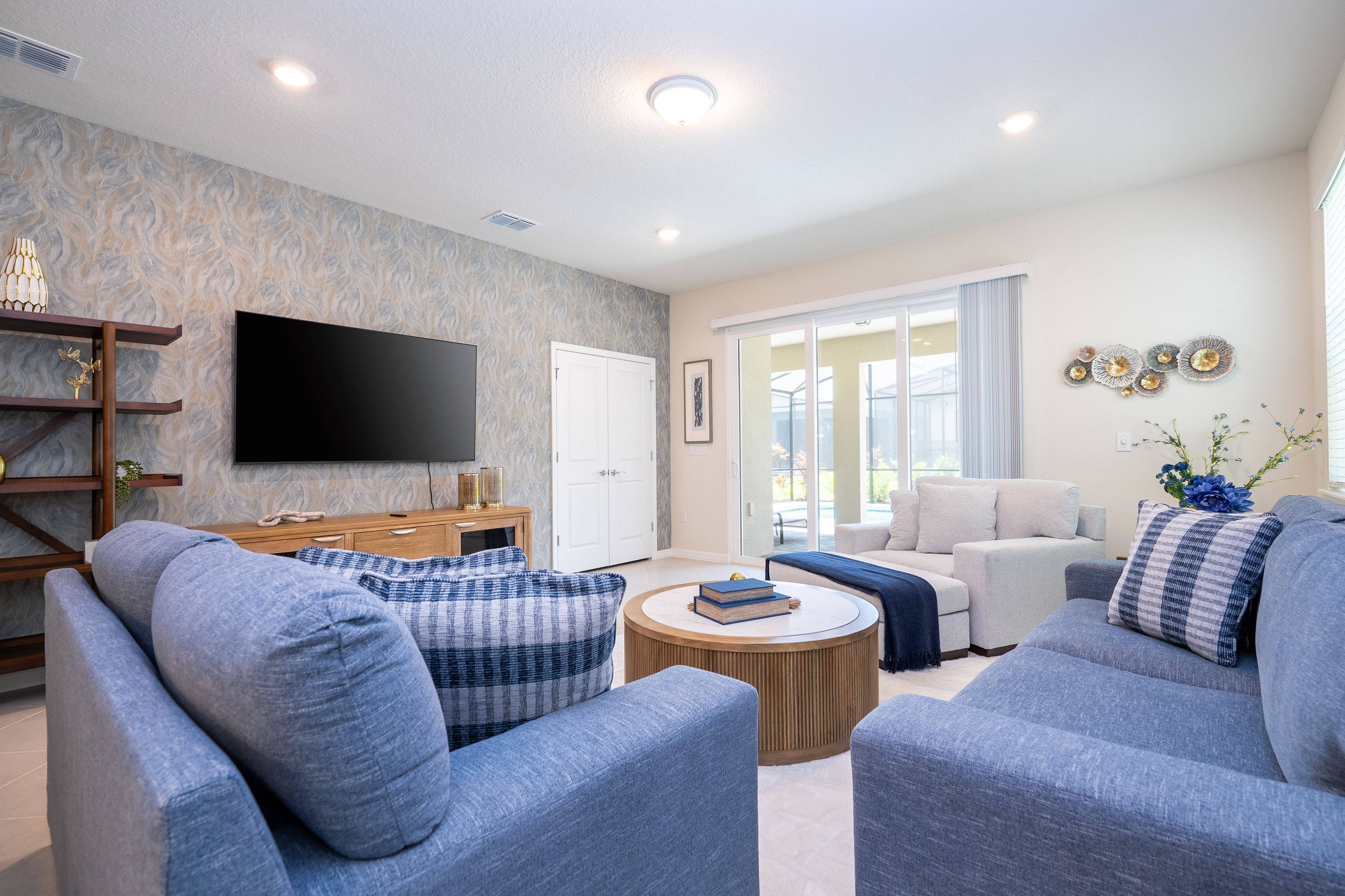 A modern living room with a flat-screen TV, grey sofas, a wooden coffee table, and a large window with curtains.