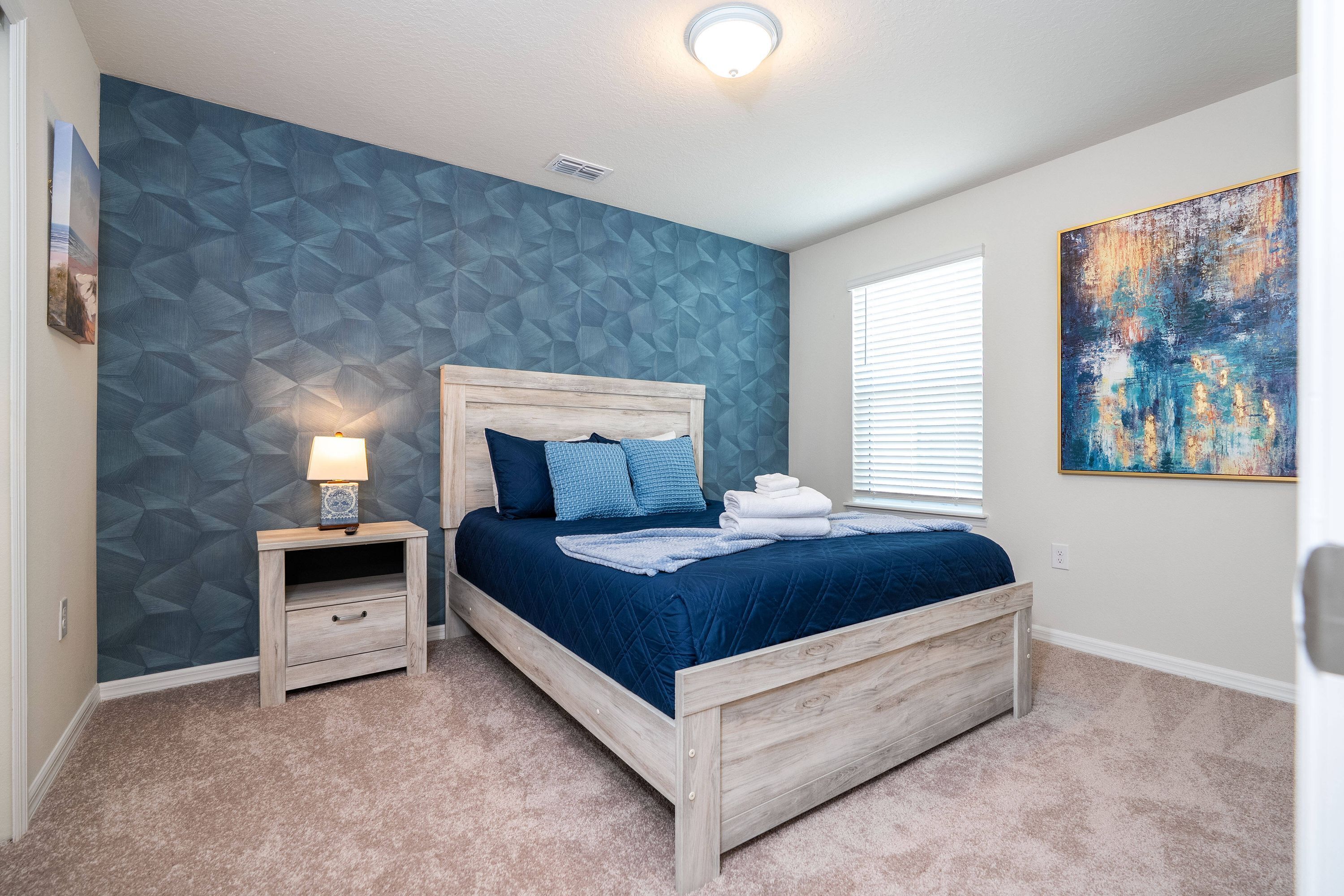 Un dormitorio con cama de madera, una mesita de noche con lámpara, pared con estampado geométrico y un cuadro abstracto grande.