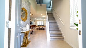 A spacious hallway with a staircase leading to a second floor. The hallway features a marble wall, a decorative wreath, and a dining area with chairs and a table. - Windsor Cay 7BR Water Park Free (Clermont)