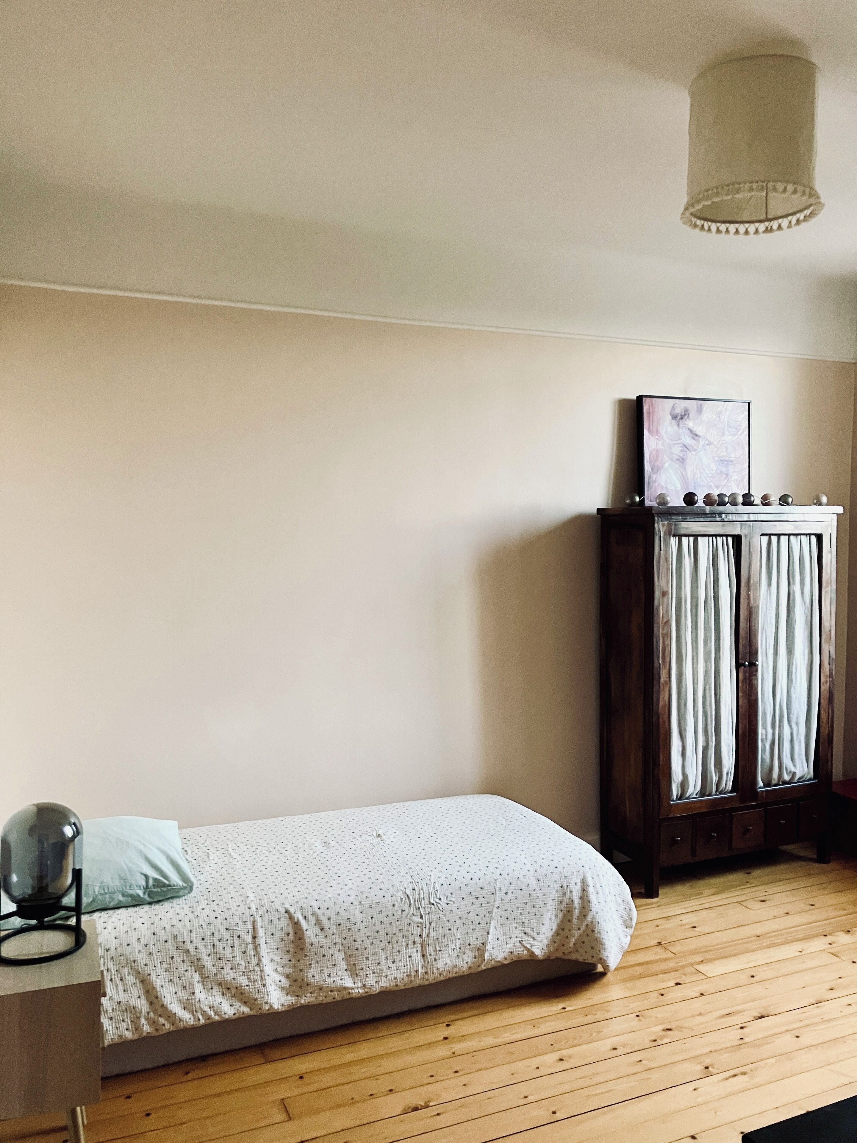 A small, single-bed room with a wooden floor, a bedside table with a lamp, and a wardrobe with curtains.