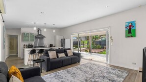 A modern living room with a grey sofa, a coffee table, and a fluffy rug. A kitchen with a stove, microwave, and cabinets is visible in the background. - Anchors Away - Mangawhai Heads Holiday Home (Mangawhai)