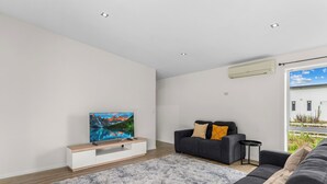 A modern living room with a flat-screen TV on a wooden TV stand, a grey sofa with cushions, a fluffy rug, and a large window with a view of greenery. - Anchors Away - Mangawhai Heads Holiday Home (Mangawhai)