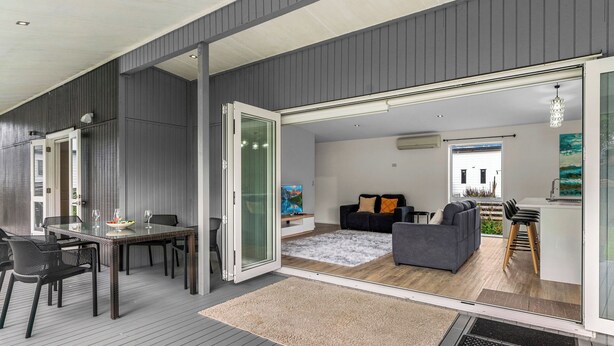 A modern living space with a dining area, a sofa, and a kitchen visible through a glass door. - Anchors Away - Mangawhai Heads Holiday Home (Mangawhai)