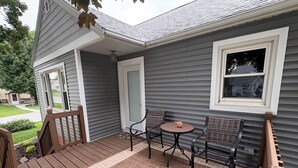 En inglasad veranda med ett litet bord och två stolar, ett trädäck och ett hus med grå träpanel och vita detaljer.
