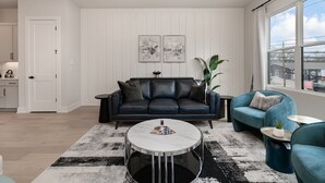 A modern living room with a black leather sofa, a round coffee table, and teal armchairs. There is a large window with a view of the city. - Trinity Beats-private Rooftop Deck-east Nashville (Nashville)