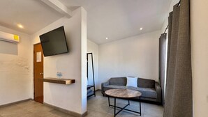 Una sala de estar moderna con un sofá gris, una mesa de centro redonda, un televisor montado en la pared y un estante de madera con botellas de agua.