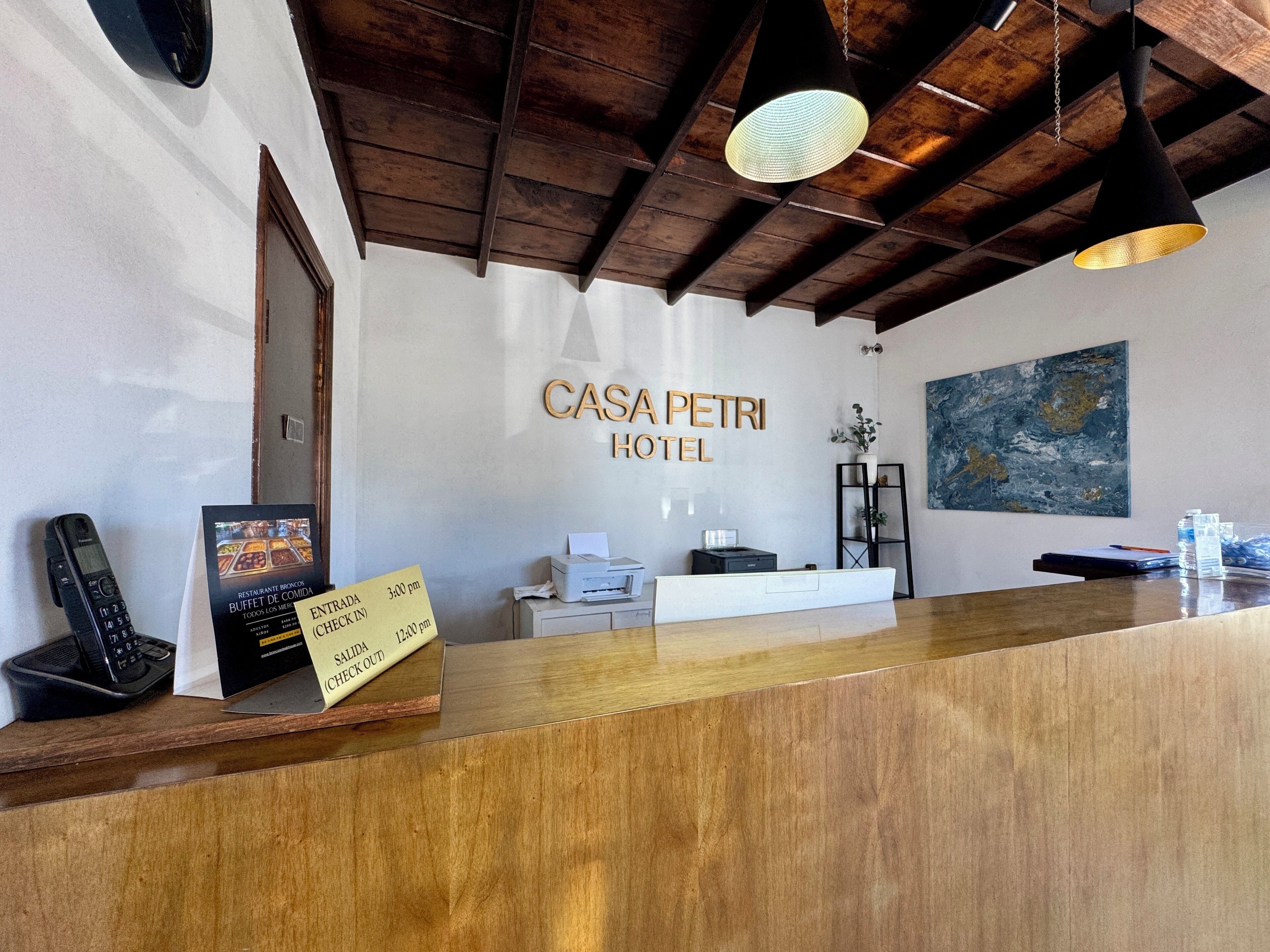 A hotel reception desk with a wooden counter, a phone, a check-in/out sign, and a painting on the wall.
