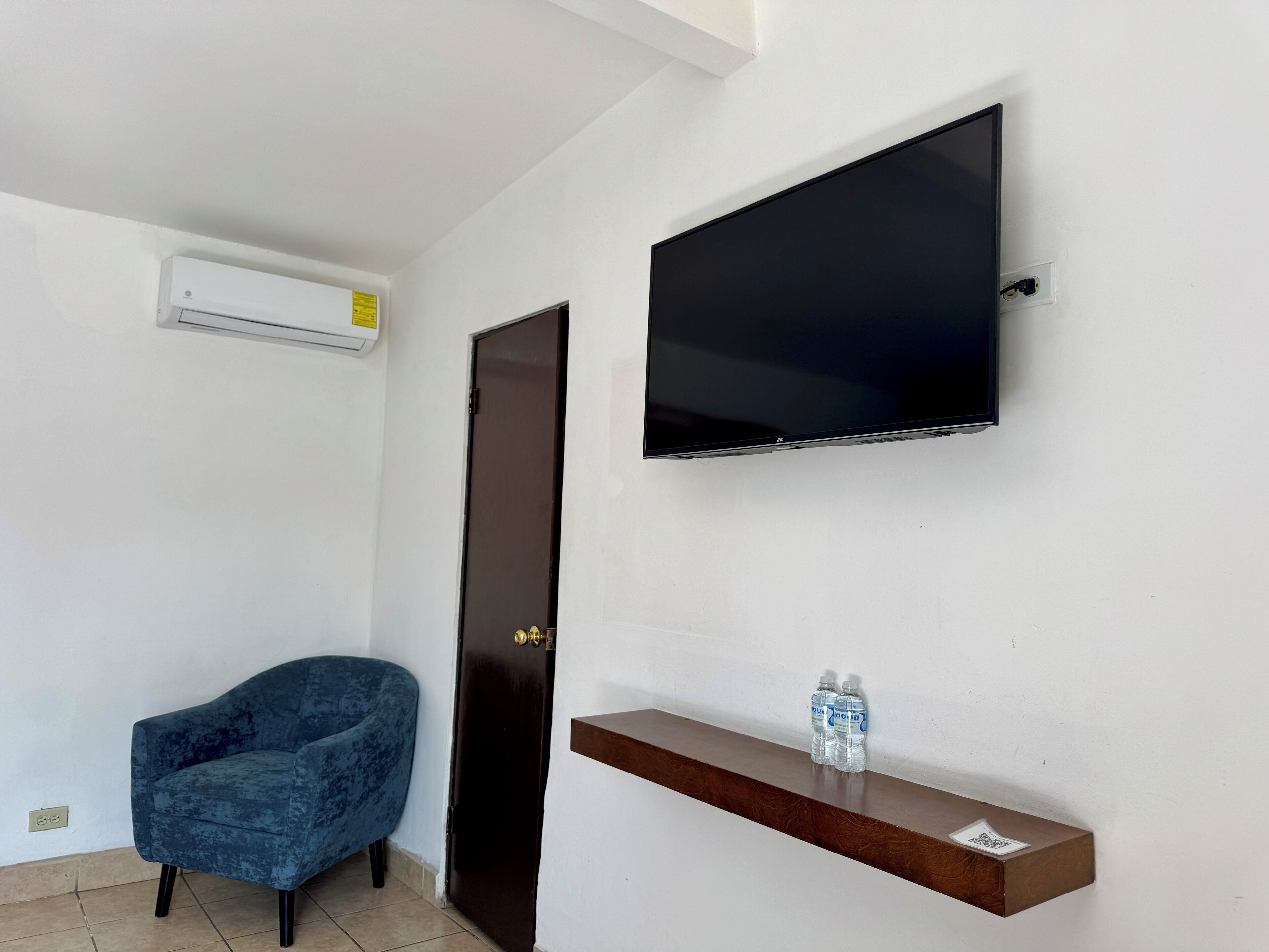 A room with a flat-screen TV mounted on the wall, a blue chair, a wooden shelf with water bottles, and an air conditioning unit.