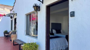 Un edificio blanco con un cartel del número 10, un balcón con un banco y plantas en macetas, y una cama con cubrecama blanco y cabecero oscuro.