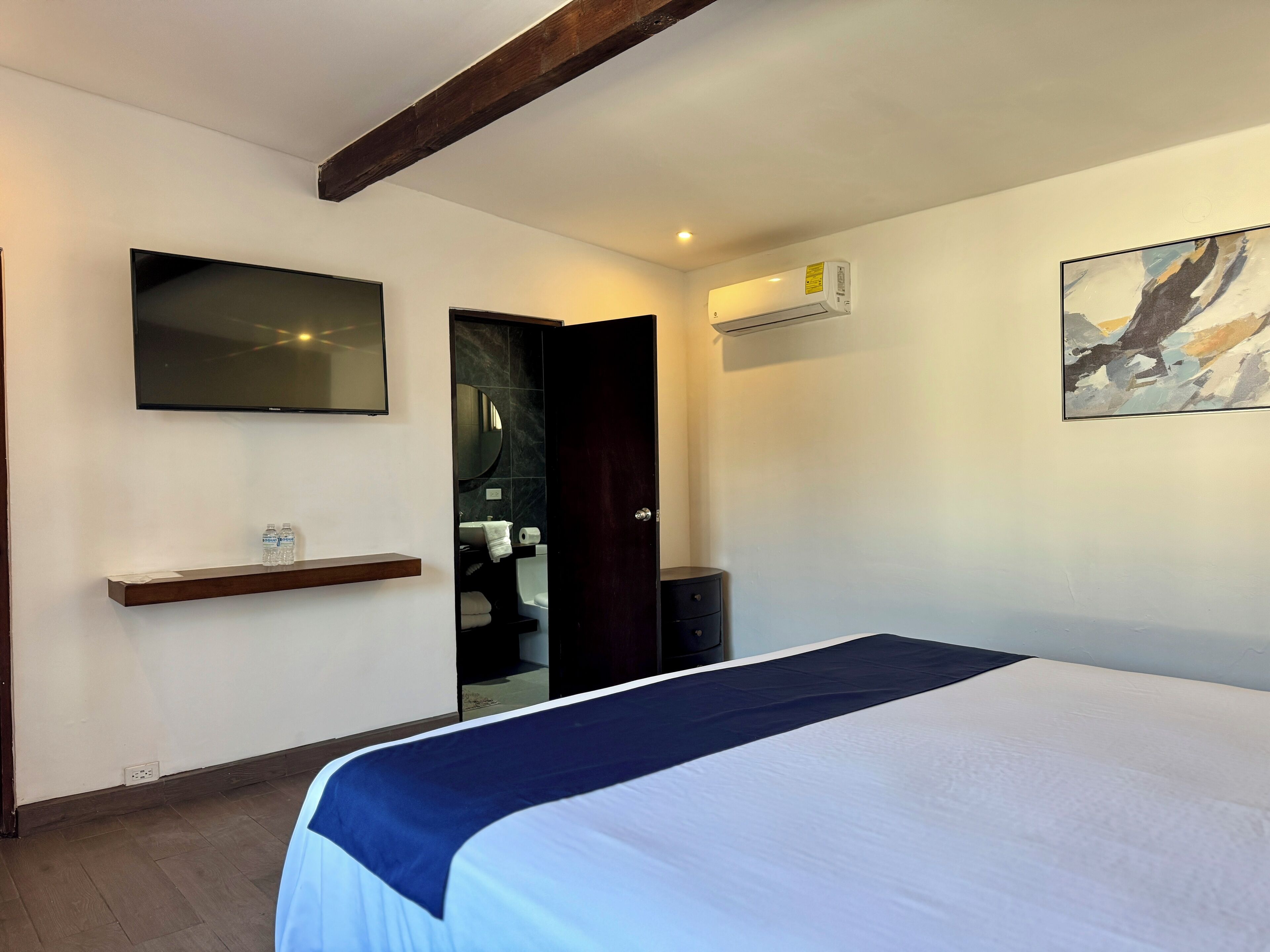 A hotel room with a bed, a TV mounted on the wall, a shelf with water bottles, a painting on the wall, and a door leading to a bathroom.