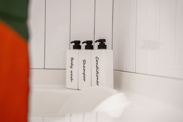 Ein Badezimmer mit weißen Fliesen und drei weißen Flaschen mit den Aufschriften 'Body Wash', 'Shampoo' und 'Conditioner' auf einer weißen Oberfläche.