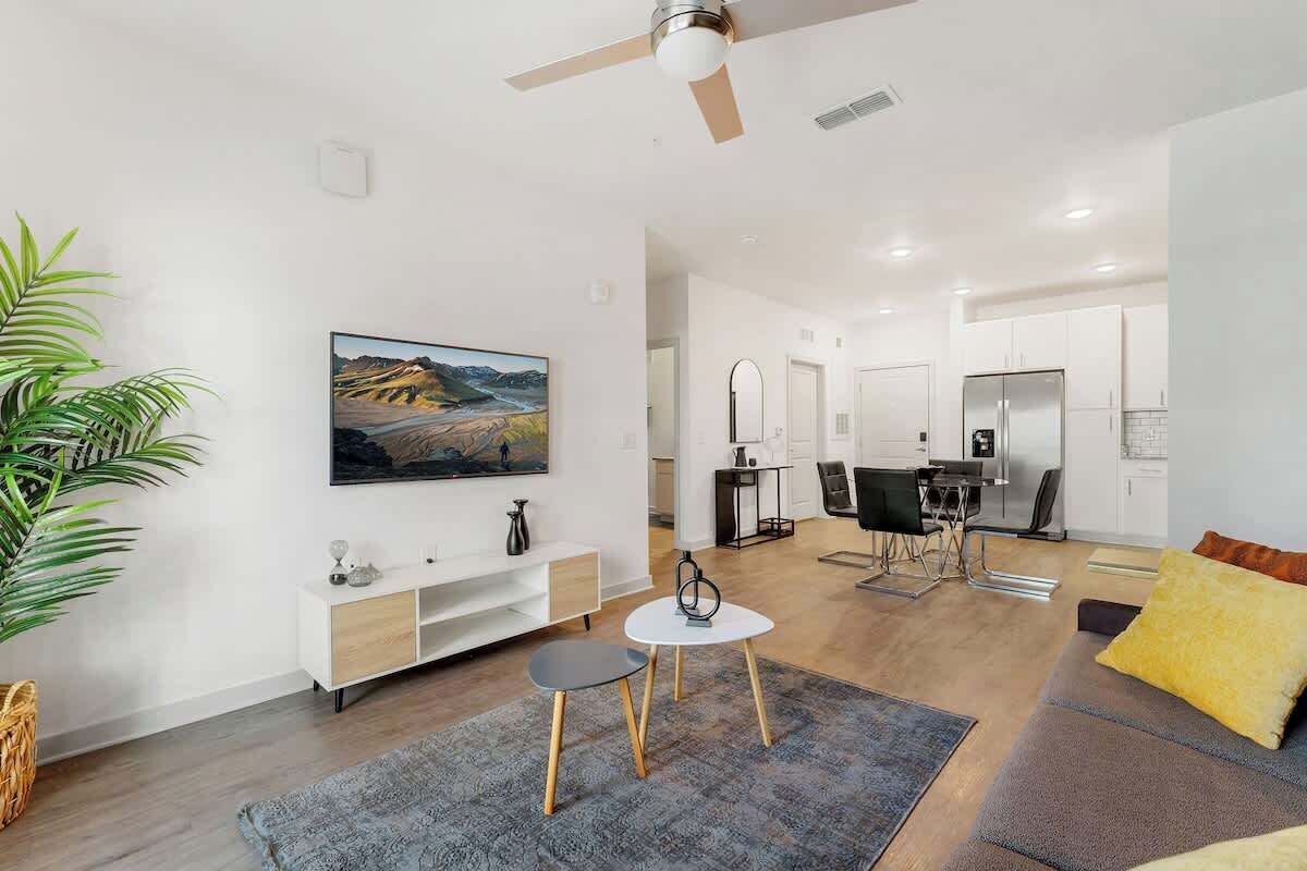 A modern living room with a flat-screen TV, a dining area, and a kitchen in the background.