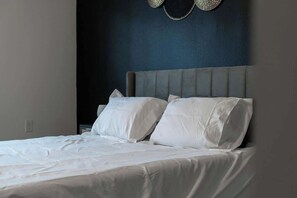 A bed with white linens and a grey upholstered headboard in a room with dark blue walls.